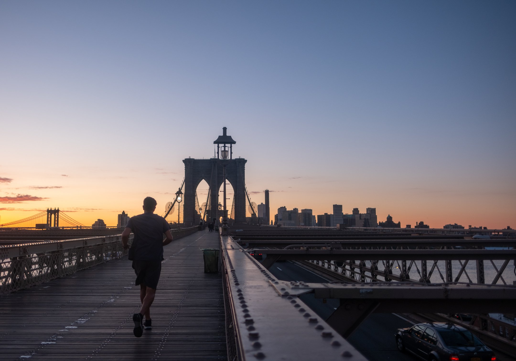 A single runner heads towards Brooklyn on the bridge, sky is orange and a few of the walkway's lights are visible
